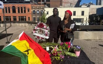 Une femme parle pendant qu'un homme se tient à côté d'elle. Un drapeau ghanéen est placé sur une table devant eux à côté de plusieurs fleurs.