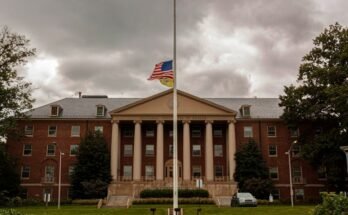 Un drapeau américain volant à moitié mât sur un mât à l'extérieur d'un large bâtiment en brique rouge avec une façade néoclassique avec `` National Institutes of Health '' écrite sur le fronton