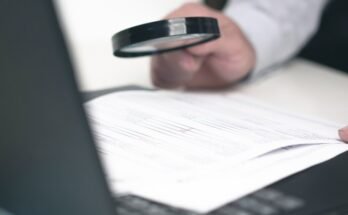 Close-up du bras pour quelqu'un qui utilise une loupe pour examiner les documents imprimés au-dessus d'un ordinateur portable ouvert.