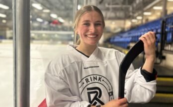 Une jeune femme vêtue d'équipement de hockey sourit à la caméra alors qu'il tient un point de hockey avec une patinoire derrière lui.