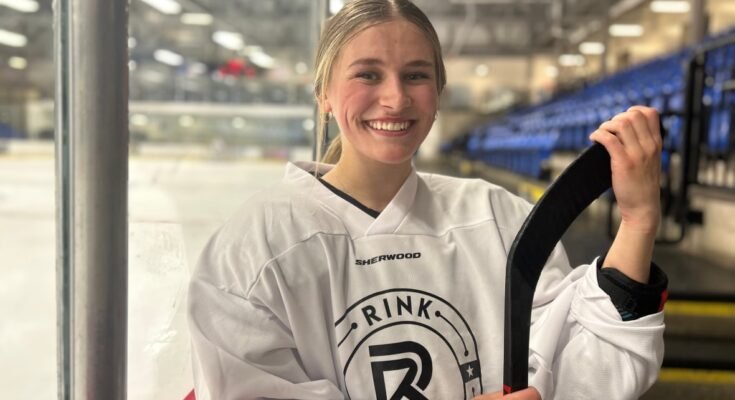 Une jeune femme vêtue d'équipement de hockey sourit à la caméra alors qu'il tient un point de hockey avec une patinoire derrière lui.