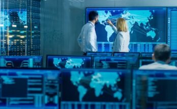 Dans la salle de surveillance du système, deux opérateurs seniors travaillent sur une grande carte interactive. L'installation est pleine d'écrans montrant des données techniques.