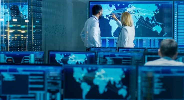 Dans la salle de surveillance du système, deux opérateurs seniors travaillent sur une grande carte interactive. L'installation est pleine d'écrans montrant des données techniques.