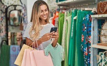 Jeune client blonde qui sourit confiant en utilisant un smartphone dans le magasin de vêtements