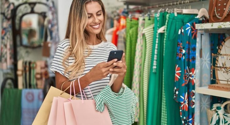 Jeune client blonde qui sourit confiant en utilisant un smartphone dans le magasin de vêtements