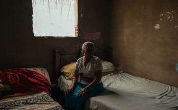 Une femme aux cheveux blancs est assis sur un lit dans une pièce sombre. Les murs ne sont pas décorés, une autre foule de lit près de qui elle se trouve.