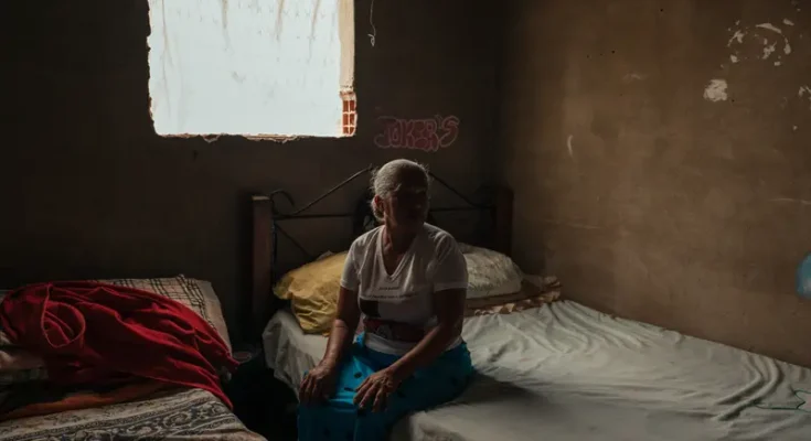 Une femme aux cheveux blancs est assis sur un lit dans une pièce sombre. Les murs ne sont pas décorés, une autre foule de lit près de qui elle se trouve.