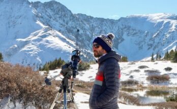 Daniel Seaton dans un chapeau de bulle en laine, entouré de montagnes à couvrage en neige en lumière du soleil debout à côté d'un instrument de type télescopique monté sur un trépied