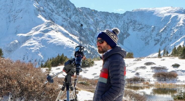 Daniel Seaton dans un chapeau de bulle en laine, entouré de montagnes à couvrage en neige en lumière du soleil debout à côté d'un instrument de type télescopique monté sur un trépied