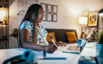 Une femme noire est assise sur son ordinateur à son bureau chez elle pendant qu'elle note