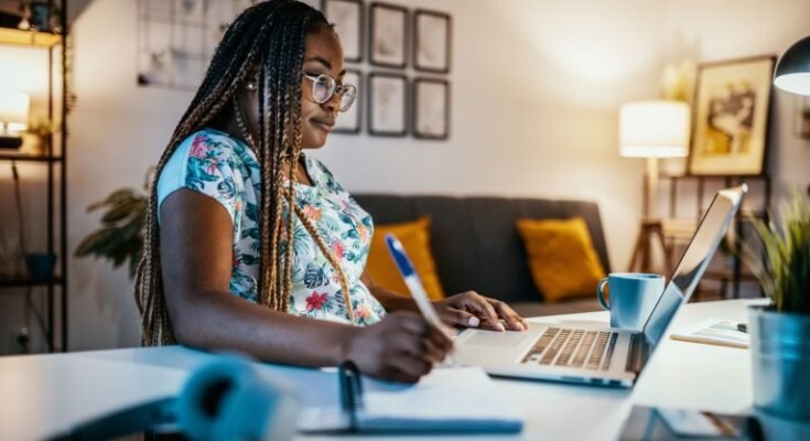 Une femme noire est assise sur son ordinateur à son bureau chez elle pendant qu'elle note